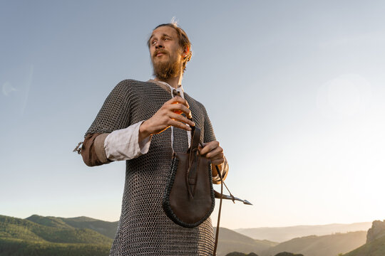 A bearded man in chainmail and tunic stands on a hilltop, gazing toward distant mountains at sunset with a leather pouch slung over his shoulder.