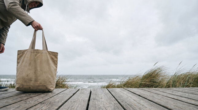 Person holding a blank reusable canvas tote bag on a weathered wooden deck by the windy cloudy coast