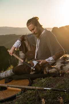 A rugged, bearded man dressed as a Viking rests on a hillside, sharpening his axe amidst scattered weaponry and golden light.