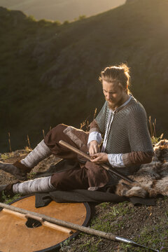 Viking warrior rests outdoors, meticulously sharpening his axe amidst a rustic landscape.