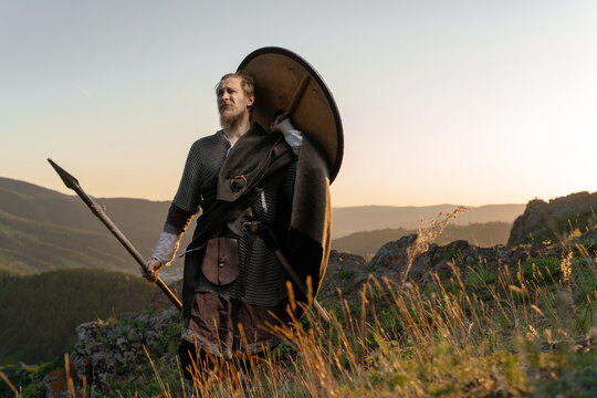 A medieval warrior stands on a hilltop, gazing out over rolling hills at sunset with shield and spear in hand.