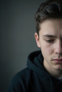 Vertical portrait of crying teenage boy with tear on his cheek. Sad young male in dark hoodie. Mental health and depression concept