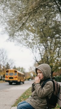 Anxious young boy sitting on bench as school bus drives away. Child biting nails and looking worried outdoors. Childhood anxiety and school concept