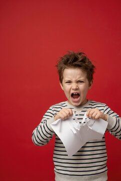 Angry young boy tearing piece of paper against red background. Frustrated child screaming and destroying homework. Emotional tantrum and stress concept. Vertical portrait