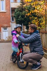 Fototapeta premium Father crouches beside his daughter on a bicycle and checks her helmet before riding. Care and protection come before every adventure