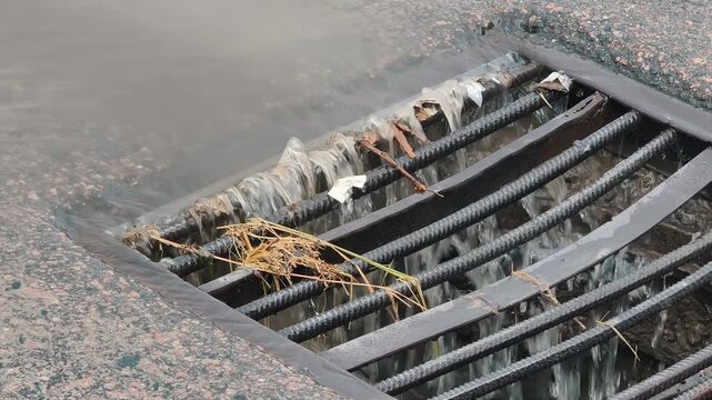 Zooming out from a close-up of stormwater runoff from a heavy rain showing water and street debris. The liquid flows rapidly down a city sewer drain through a metal grate.