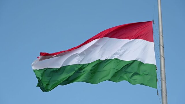 Hungarian national flag flying high and waving in the wind. red, white and green stripes. shiny silk fabric. blue sky background. metal flag pole. patriotism concept. national symbol. 