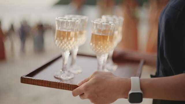 A waiter carrying a wooden tray with elegant crystal champagne flutes filled with sparkling wine, serving guests at a luxury outdoor wedding reception or upscale corporate party.