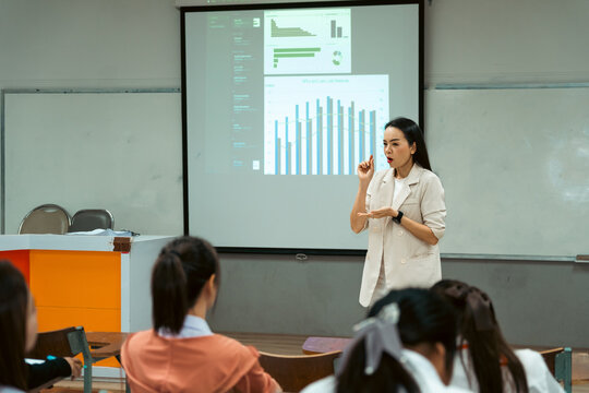 Female professor delivering an engaging academic lecture with crisp data charts on a projector screen to focused university students in a modern classroom