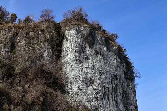 Grey stone cliff overlooking the landscape near Arona on Lake Maggiore. Leafless trees, green grass and bushes under a clear blue sky on a sunny spring day. Natural rocky landscape in northern Italy.