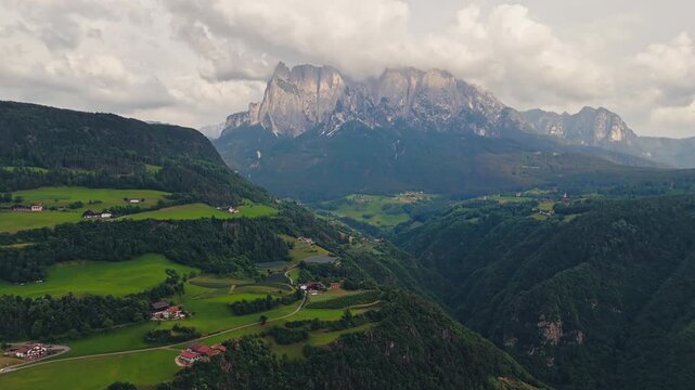 Rolling green hills and forest patches with small settlement below Dolomite mountains, aerial view