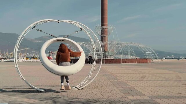 Unrecognizable young female in warm fuzzy coat enjoying dynamic swinging motion on modern round public installation at Novorossiysk waterfront with commercial port and sunny mountain background