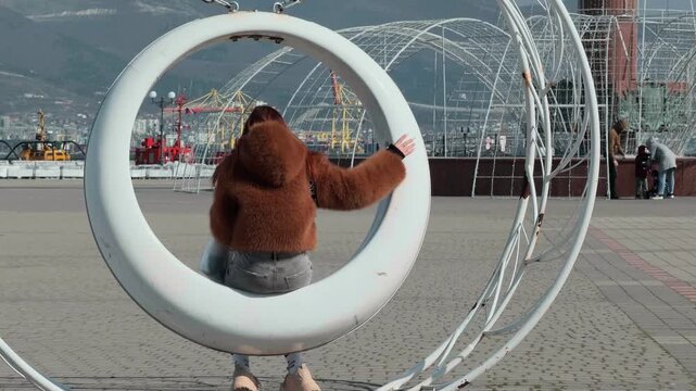 Unrecognizable young woman in fuzzy jacket relaxing on modern circular swing at city square in Novorossiysk overlooking seaport cranes and distant mountains during bright sunny day authentic rear view