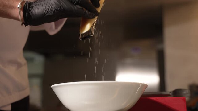 Professional chef in black gloves grating hard cheese into a white bowl in a restaurant kitchen, fa01