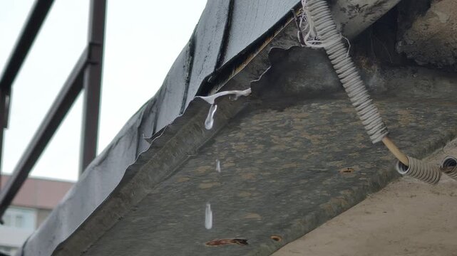 Water drops fall rapidly from the jagged edge of a rusty corrugated metal roof during a rainy day. Concept of seasonal change, spring thaw, and deteriorating urban infrastructure.
