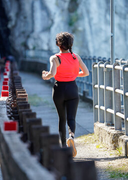 Female runner training alone on mountain road path beside railing and rock wall. Rear view shows active lifestyle, endurance and quiet outdoor fitness