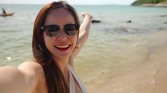 Close-up selfie portrait of a beautiful young Asian woman wearing sunglasses on a tropical beach, smiling and waving her hand at the camera. Happy female tourist taking a selfie and enjoying a summer.