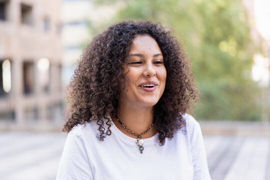 Young woman laughing softly during an outdoor urban conversation. Gen z lifestyle portrait with curly hair and bright natural mood. Joyful wellbeing concept