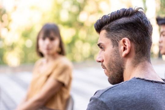 Serious young man in profile during a tense outdoor conversation. Blurred friends sit nearby at a patio table with soft bokeh. Relationship conflict concept