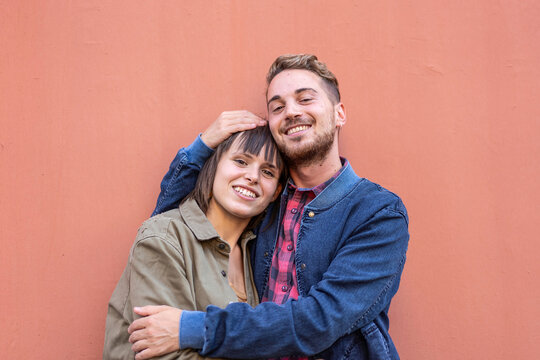 Happy young couple hugging against a coral wall in a warm lifestyle portrait. Gen z or millennial friends share affection and tenderness outdoors. Relationship concept