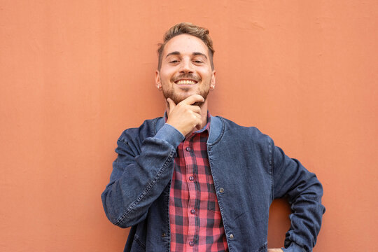 Confident young man smiling with hand on chin against a warm coral wall. Casual gen z or millennial portrait with denim jacket and playful charisma. Personal branding concept