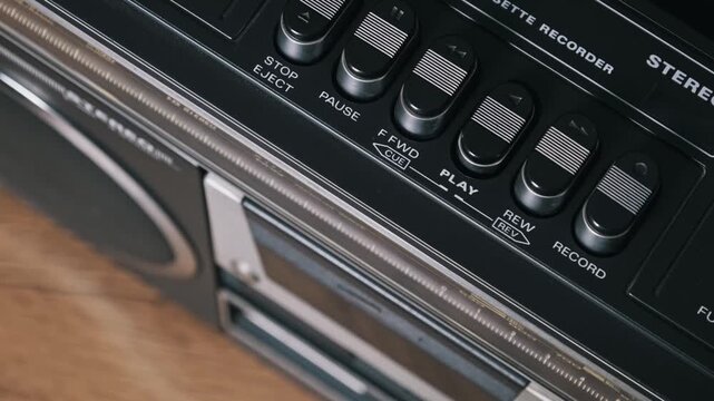 High-angle close-up of a finger pressing the play button on a black retro stereo. The wooden table and white text labels highlight the vintage 80s nostalgia.