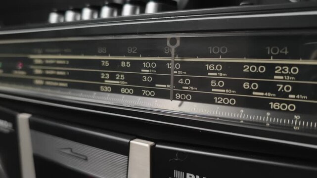 Angled shot of a vintage boombox radio tuner needle scanning the frequency display. The dusty scale and wave bands emphasize the classic design of the portable player.