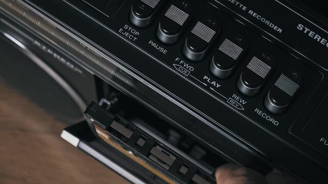 Close-up of a hand inserting and playing an old audio cassette in a vintage black stereo. The worn yellow label and chrome details highlight the retro music experience.