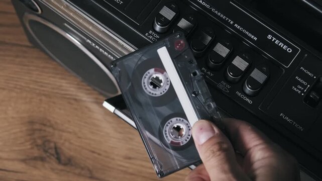 High angle shot of a hand ejecting and flipping a transparent cassette tape in a vintage recorder. The chrome details and wooden surface highlight the retro music hobby.