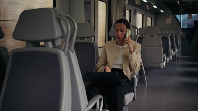 A young businesswoman works on her laptop and talks on her smartphone while commuting on a modern train. She is multitasking during her journey.
