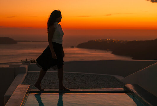 Golden hour serenity &mdash; a woman&rsquo;s silhouette walking beside a pool overlooking Santorini&rsquo;s glowing caldera at sunset.