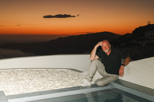 A man sits beside a pool on a modern terrace overlooking the glowing caldera of Santorini, Greece, during sunset. The warm orange sky and tranquil sea create a serene, luxurious travel moment.
