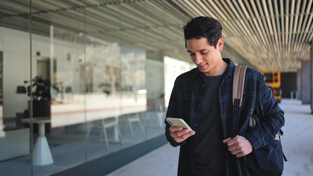 Young hispanic man walking while using mobile phone. Happy university student in the city