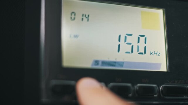 A finger presses buttons on a multi-band radio switching between medium wave and FM modes on the digital LCD. The frequency numbers change as the receiver scans for a station.