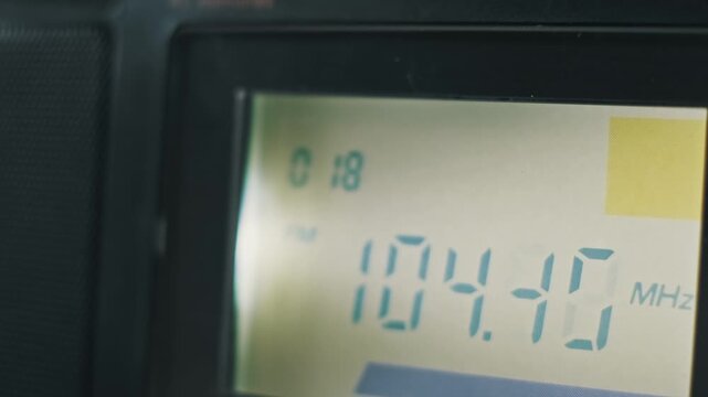 Extreme close-up of a digital radio LCD screen auto scanning upward on the FM band. A finger holds the scan button as teal frequency numbers in MHz rapidly increase.