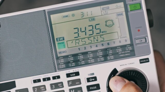 Top-down view of a person tuning a portable shortwave radio. A finger adjusts the manual dial changing the SW frequency on the LCD screen which shows digital station information.