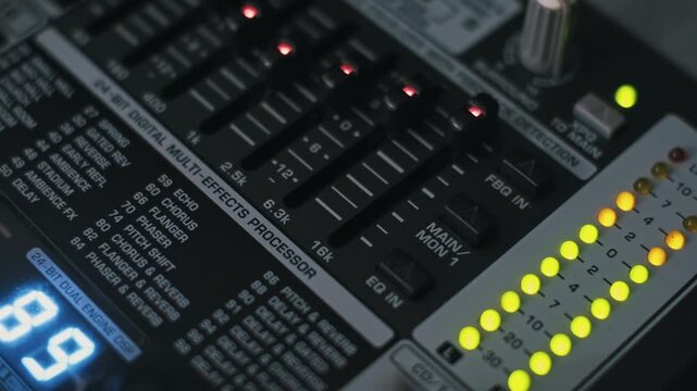 Angled close-up of a digital multi-effects processor on an audio console. The shot pans to reveal EQ faders, active yellow LED meters, and a blue digital display in a studio.