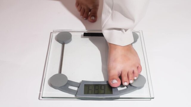 Woman&rsquo;s legs in white pants step onto a bathroom scale on a clean white background. Close-up of feet with a light gel pedicure standing on digital scales. Weight control, weight loss journey, healthy 