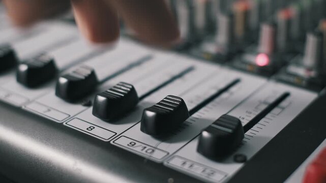 Angled macro view of fingers pulling down two faders on a professional sound mixing board. The action demonstrates lowering volume near a decibel scale and numbered channels in a studio.