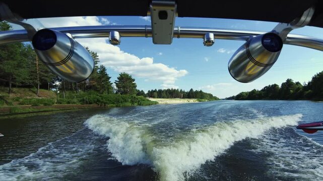 A wide cinematic symmetric shot from a speed boat, showing a large white water wake on a river, framed by large chrome wakeboard tower speakers under a blue sunny sky