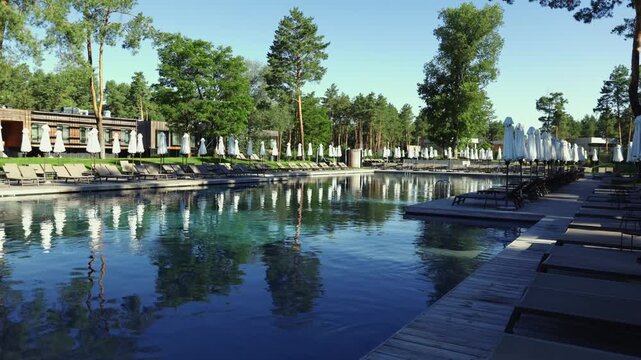A wide cinematic establishing shot of a large swimming pool at a luxury resort, lined with rows of sunbeds and closed white umbrellas, surrounded by tall pine trees on a bright sunny day