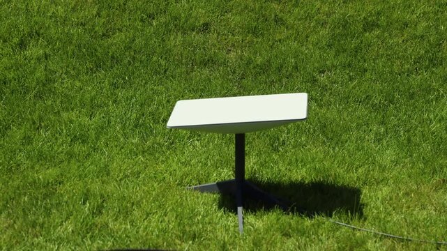 A wide cinematic shot of a white Starlink satellite internet antenna dish setup on a vibrant green grass lawn on a bright sunny day