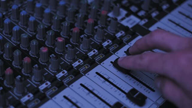 Close-up of a hand adjusting faders and pressing mute on an audio mixing console. The equipment is illuminated by shifting purple, blue, and red club lights for a DJ live event.