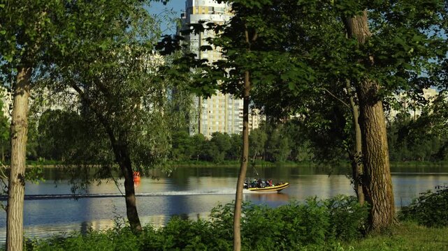 A wide shot of a yellow rescue boat speeding on a calm river with passengers on board, framed by green trees with city residential high-rise buildings in the background