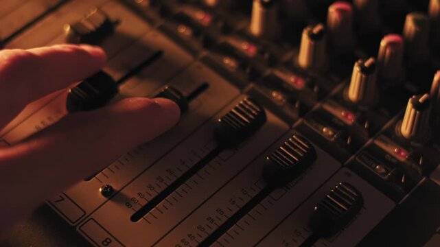 Close-up of audio mixer faders on a professional soundboard showing volume level markers and textured knobs. Shifting purple, blue, and orange club lights create a dynamic atmosphere.