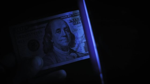 A hand checks a 100 dollar bill with UV light, revealing glowing security strips in the dark. Concept of counterfeit detection, banking security, and financial fraud prevention.