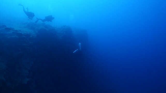 scuba divers exploring around a reef underwater deep blue water big rocks and bubbles ocean scenery 