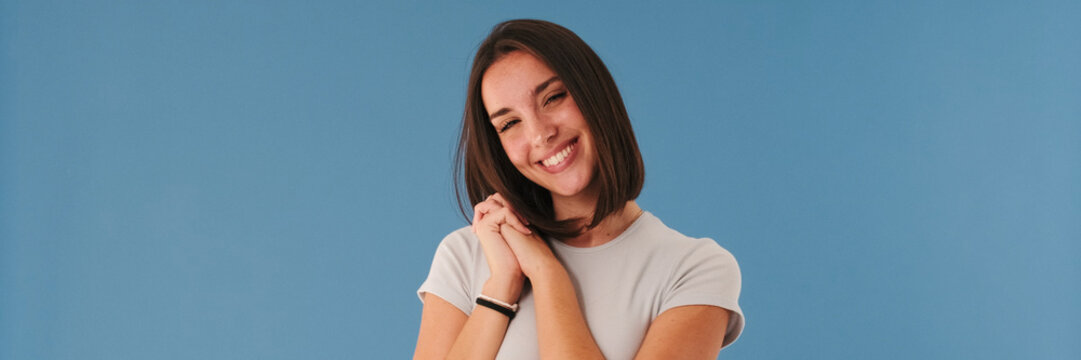 Smiling beautiful young woman brunette dressed in white top looking at camera folding her palms so cute isolated on blue background in studio. Panoramic