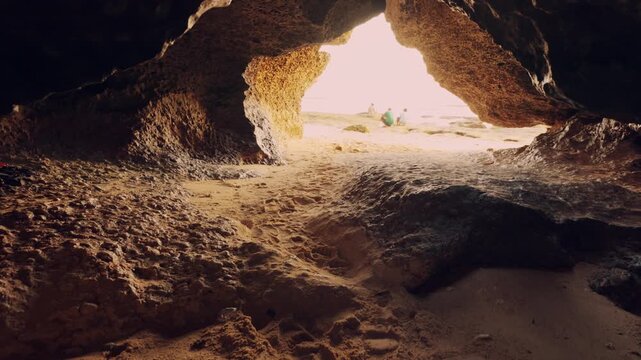 Low angle shot focusing on the sandy texture of a cave floor leading towards the blinding light of the beach entrance. Natural rock architecture framing a summer day.
