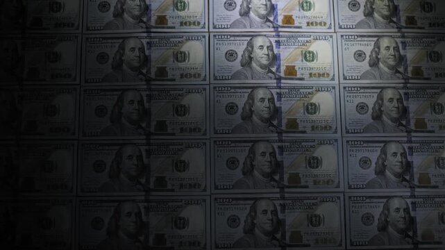 Top-down view of a silhouetted hand using a handheld UV light to scan columns of hundred dollar bills. The inspection verifies banknote authenticity by revealing hidden security features.